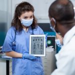 Pediatrician nurse with protective face mask against covid19 holding tablet computer with bones radiography on screen. African american doctor explaining medical expertise in hospital office